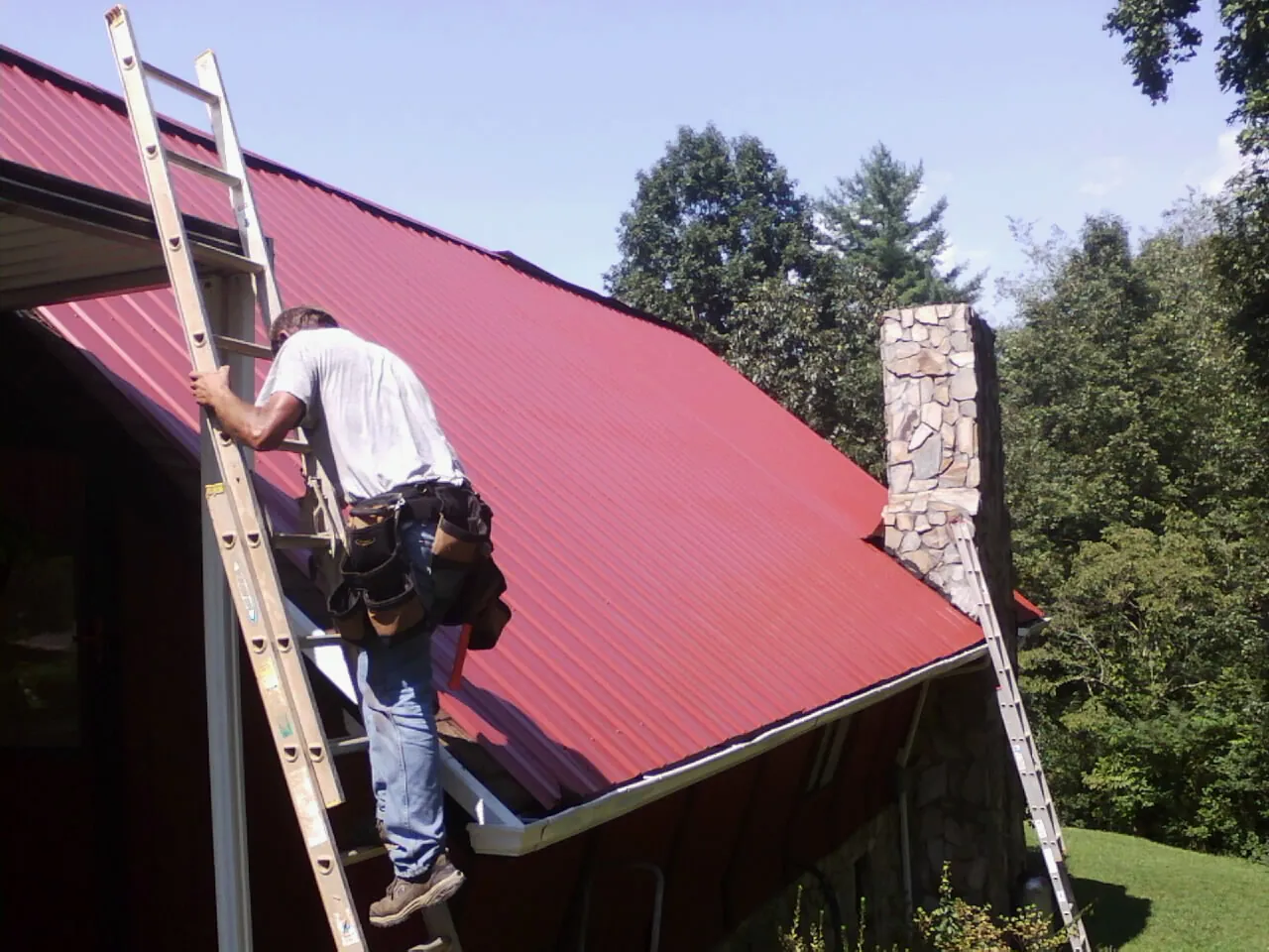 Roofer on a ladder beside a red metal roof and stone chimney — Soffit & Fascia Repair by Roofing Brothers in Fredonia